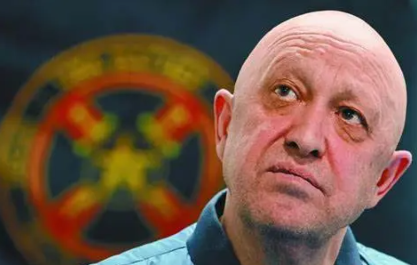 A bald Yevgeny Prigozhin tilts his head, raises his gaze to the right, and looks pensive. Behind him is some sort of red, green, and yellow logo, although it is too blurred to see clearly.