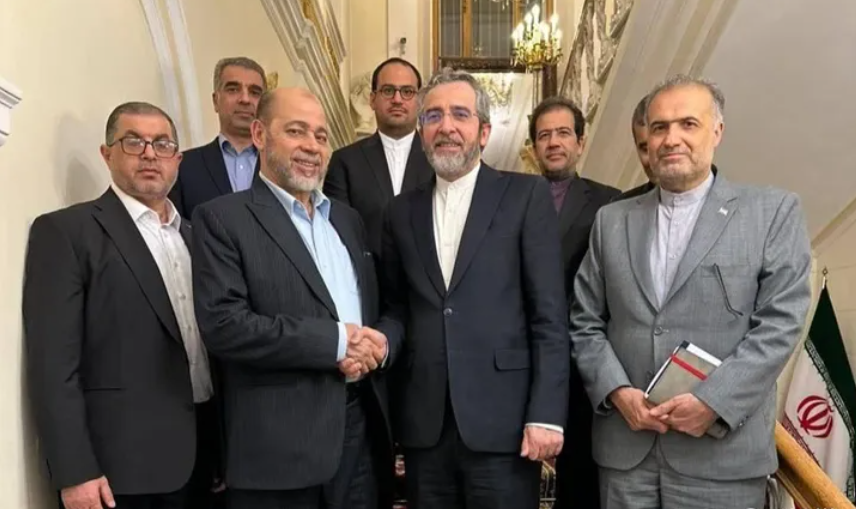 A group of seven bearded men in suits pose on a staircase, as the two men in front smile and shake hands.