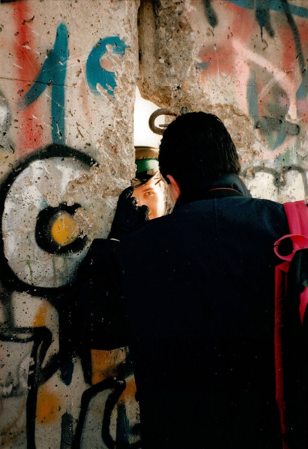 Through a large gap in a spray-painted section of the concrete Berlin Wall, a West German man with a black coat and gloves and a red backpack speaks to a uniformed East German guard wearing a khaki military cap.
