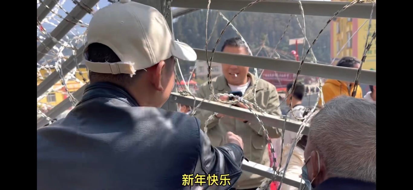 A young man wearing a black leather jacket and tan baseball cap, accompanied by an older man wearing a surgical mask, reaches a hand through metal bars and barbed wire. A number of people are visible on the other side of the bars, including a man wearing a tan worker’s jacket and holding a cell phone.