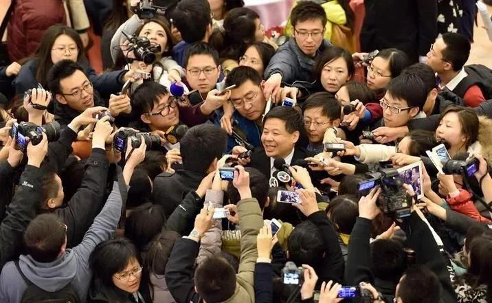 A government official is surrounded by dozens of reporters waving microphones, miniature recording devices, and digital cameras.
