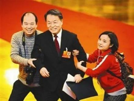 A government official in a suit and tie smiles awkwardly as Zhao Qing (clad in a red zip-up jacket and backpack) hangs on to his left arm, while a male photographer (wearing a gray sweater, with a large camera around his neck) clings to his right arm.