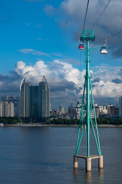 Red and white cable cars, suspended high in the sky, pass an enormous turquoise-painted steel support structure as they cross a river in Harbin. In the background is a brilliant blue sky, dramatic cloud formations and a forest of skyscrapers.