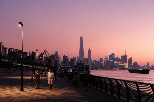 A couple walks along a cobbled street alongside a river in Shanghai at sunset. Enormous skyscrapers are silhouetted against the pale pink sunset, and the surface of the river reflects the rays of the setting sun.