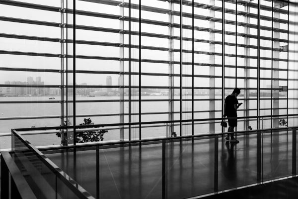 Black and white image of a passenger walking in an empty airport terminal, against floor to ceiling windows that look out on the ocean.
