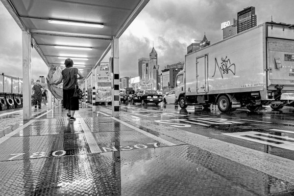 Black and white image of a woman with an umbrella in a covered, lighted walkway in Wan Chai, Hong Kong, as traffic passes in the street beside her, and light reflects from the water on the pavement.