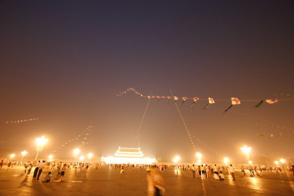 Kites fly overhead as crowds of people sit and stand in Tiananmen Square, Beijing, at night. Lamplights illuminate the scene, and in the background, Tiananmen Square is ablaze with lights.