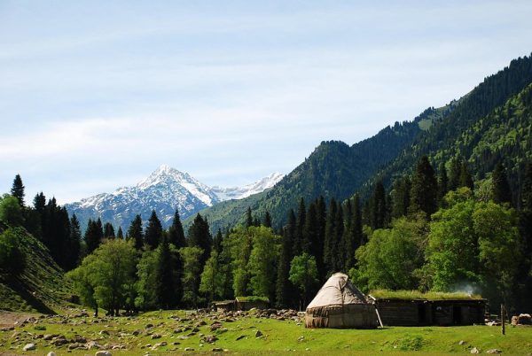 A brown yurt nestles in a lush green valley, with towering snow-capped peaks in the background. This photo was taken in Ili Kazakh Autonomous Prefecture, northern Xinjiang, China.
