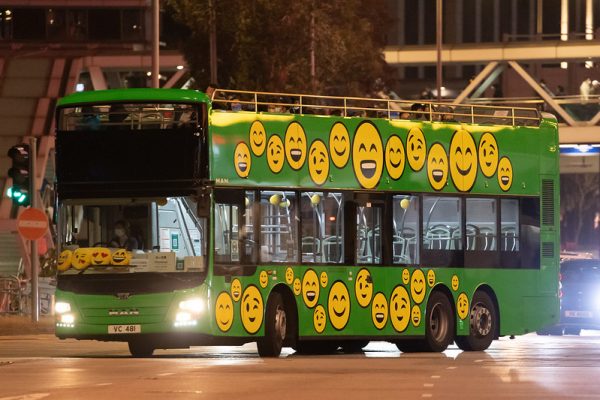 A green double-decker sightseeing bus in Hong Kong is decorated with dozens of cheerful yellow smiley-face emojis.
