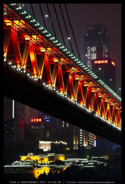 Red and yellow lights illuminate the criss-crossed metal girders of a bridge, prominent in the foreground, against a backdrop of brightly lit skyscrapers and red neon signs.