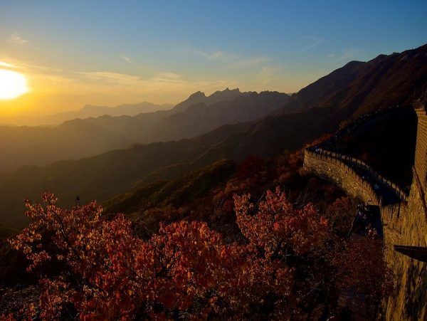 Yellow rays of the setting sun illuminate the rolling hills and red autumn leaves near a portion of the Great Wall.