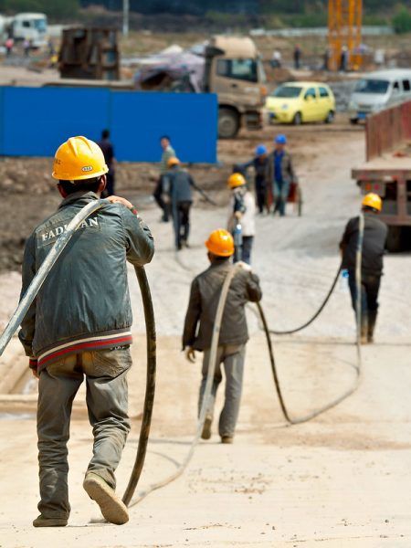 A line of five workers in yellow hard hats, their backs to the camera, carry a long, thick black cable on a construction site in the city of Kunming, Yunnan Province, China. The worker in the foreground is seen in sharp detail, whereas the other workers and the construction site in the background are somewhat blurred.