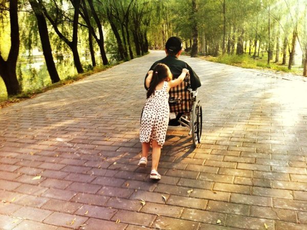 Moving away from the camera, a young girl with a ponytail pushes an older woman in a wheelchair down a cobbled pathway lined with pale green willow trees and illuminated with dappled sunlight.