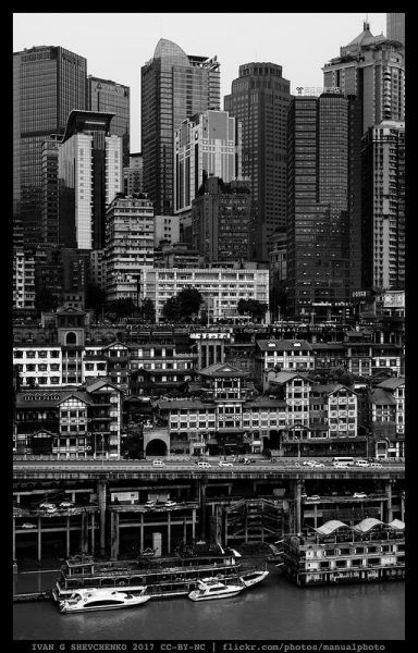 Black and white boats and docks fill the foreground of this image of Chongqing’s riverfront, giving way to densely packed three- and four-story buildings and warehouses, and a forest of gleaming glass high-rises rising behind them.
