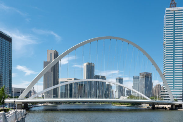 Two dramatic arcs of Tianjin’s tiered-arch Dagu bridge stretch across a slate-colored river, against a backdrop of gray and silver highrises and a bright blue sky with wispy clouds.