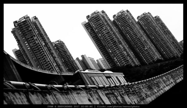 Tilted black and white photo of a grey urban landscape, with bridge in the foreground and towering apartment blocks in the background.