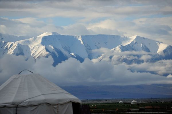 In the foreground, a white yurt looks out on a green wetland dotted with other yurts, and beyond that, towering snow-capped peaks wreathed with low clouds.