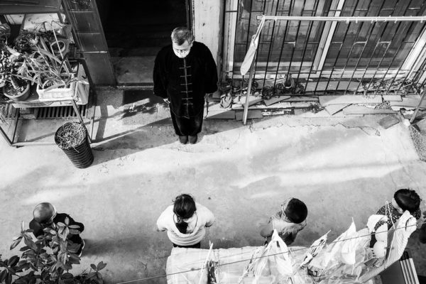 Viewed from above, a man wearing a black traditional Chinese gown and a white surgical mask stands outside a building facing a line of four children, also wearing masks.