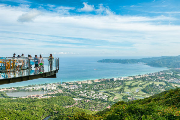 At left, a group of tourists on a glass-fenced circular walkway appear to float in the sky at a dizzying height, far above the spectacular vista of Sanya Bay on the island of Hainan, China.