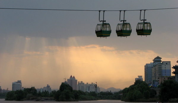 Three cableway cars, suspended from a cable high above the Lanzhou cityscape, look dark against the rays of the setting sun spilling through the clouds.