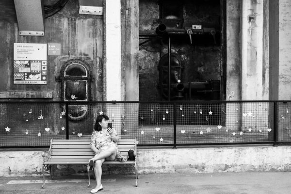 A bare-legged woman in a summer dress sits on a bench, with one ankle propped on a knee, holding her cell phone and looking askance at something off-camera. Behind her is a fence in Qingdao adorned with “lovers’ locks.”