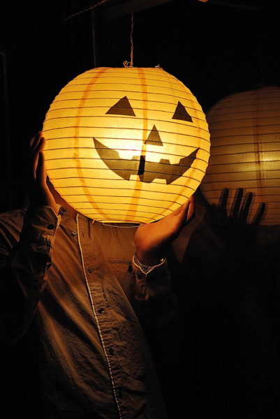 A round orange lantern with a jack-o'-lantern face glows against the darkness, and obscures the face of the person holding it.