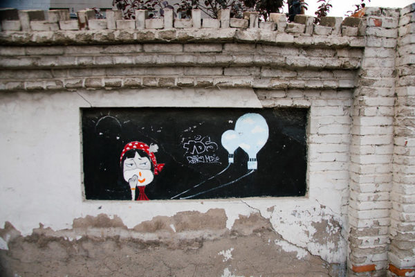 Street art painted on a crumbling gray-and-white brick wall depicts a cartoon female face with a red-and-white polka-dotted scarf smiling at two blue hot air balloons, against a black background.
