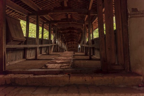 Interior view from one end of an old wooden covered bridge. The dark wood arches into the distance, green trees are visible on either side, and a lone person stands within the bridge’s corridor and gazes out at the scenery.