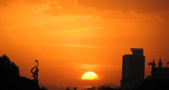 Against a vivid orange sunset and an enormous yellow setting sun wreathed with orange clouds, the trees and buildings of Lanzhou (the capital of Gansu Province, China) are but dark silhouettes.