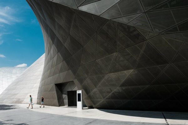 Gleaming gray panels adorn the sweeping facade of the futuristic Museum of Contemporary Art in Shenzhen. Two people are walking out the front door of the museum, and a hint of blue sky and puffy white clouds is visible in the top left corner.