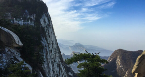 A stunning view of a monastery perched on the edge of a sheer cliff, dramatic pine trees, and verdant hills shading off into the distance.