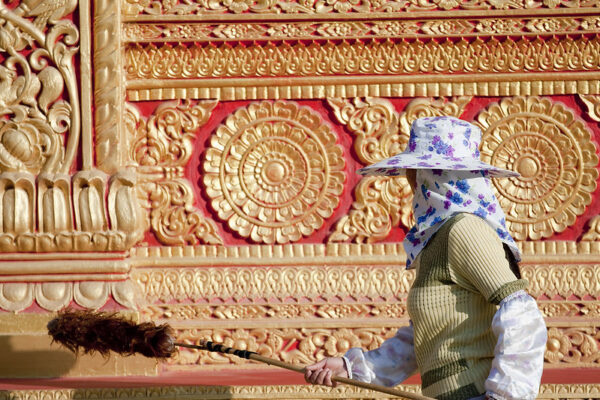 A woman wearing puffy sleeve-protectors and a flowered sun-hat and neck-protector wields a long wooden feather duster to clean the vermillion-and-gold facade of a temple in Yunnan Province, China.