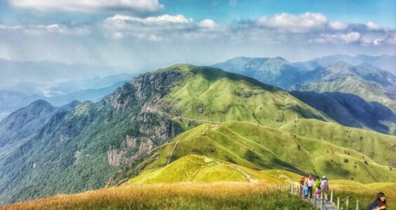 Against a backdrop of teal skies and wispy clouds, a small group of hikers walk along a narrow path atop the rolling, verdant hills of the Wugong mountain range in Jiangxi province, China.