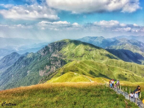 Against a backdrop of teal skies and wispy clouds, a small group of hikers walk along a narrow path atop the rolling, verdant hills of the Wugong mountain range in Jiangxi province, China.