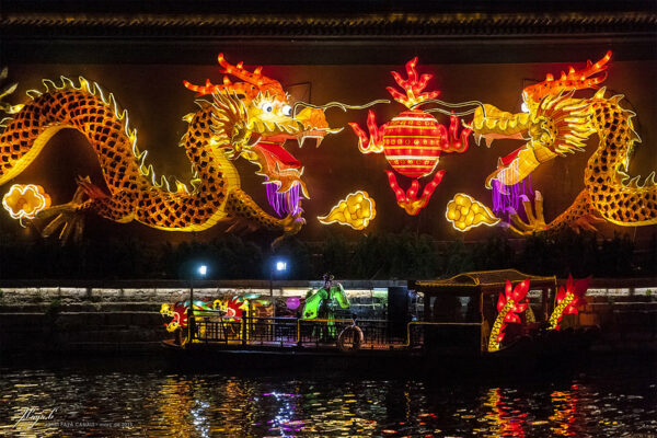 A female dancer in an elaborate headdress and bright green traditional Chinese gown performs on a barge adorned with brightly illuminated dragons, against a backdrop of two even larger and more brilliantly lighted dragons and a large red lanterns. The light from the Chinese New Year decorations is reflected in the dark water below. 