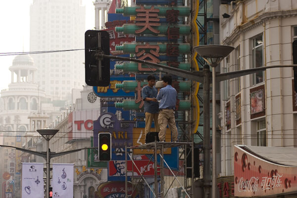 Two maintenance men in blue shirts and khaki slacks stand atop scaffolding to reach a surveillance camera mounted near an intersection of Shanghai’s bustling Nanjing Road.