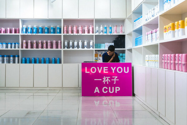 In a brightly-lit store linked with shelves of colorful thermoses and cups, a young man in glasses and a navy t-shirt sits behind a large pink sign that reads “LOVE YOU 一杯子 A CUP,” in which “a cup” (yi beizi) is a sound-alike for the phrase “my whole life long.” 