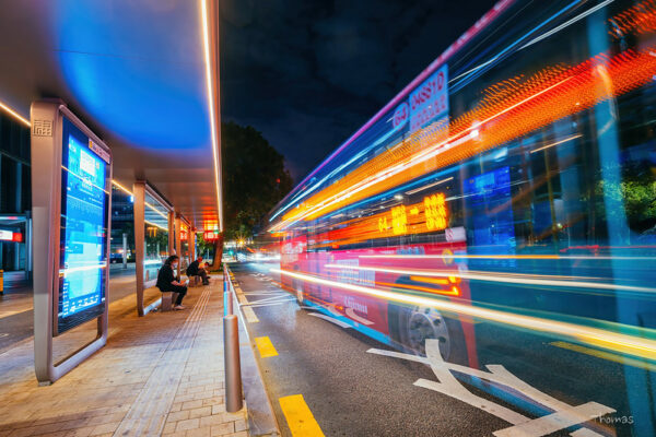 Streaks of yellow, orange, red, and blue light inform this stylized photo of a bus speeding by a bus stop at night in Shenzhen, as two passengers wait on benches.