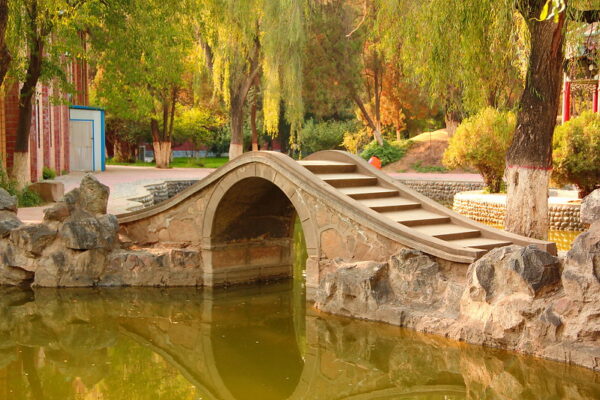 A small body of water, an arched stone bridge, green willow trees, and brick buildings adorn the campus of Lanzhou University.
