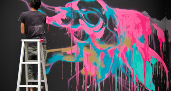 A short-haired artist in red tennis shoes standing on a ladder uses neon pink, blue, and turquoise spray paint to create a surreal elephant-like image on a black wall.