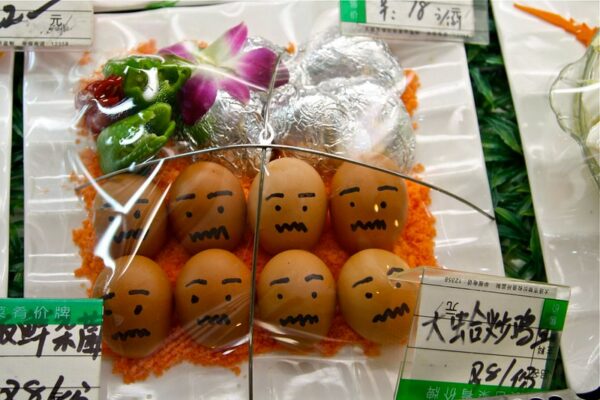 Someone has used black felt-tipped marker to draw eyes, eyebrows, and squiggly mouths on eight brown eggs, wrapped in plastic for sale with some accompanying vegetables.
