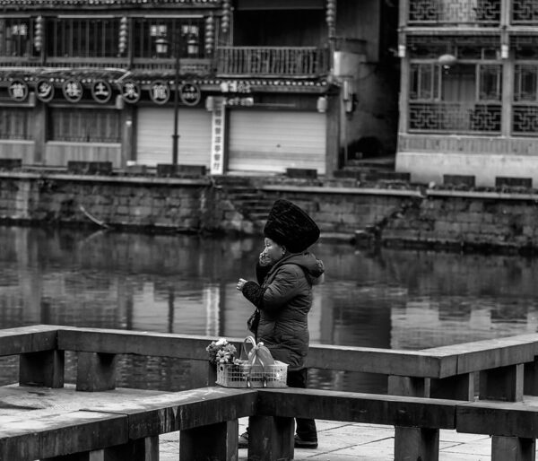 A woman with a puffy jacket, basket of flowers, and a large traditional headdress talks on a cell phone as she walks alongside a canal lined with charming traditional buildings in the ancient town of Fenghuang, Hunan Province. 