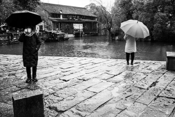 In a rainy landscape of cobblestones, tile-roofed houses, and a canal lined with trees and low-slung houseboats, two figures dressed in coats and carrying umbrellas face in opposite directions. The woman with a black coat, black umbrella, and white face mask faces the camera, while another person with a white coat and white umbrella faces in the opposite direction, gazing at the canal.