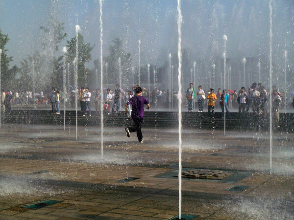 A young man in a purple t-shirt, black pants, and sneakers races through a public square from which dozens of fountains, placed every few yards, spout streams of water high into the air. Groups of people stand around the perimeter of the fountains, watching the fun while staying dry.