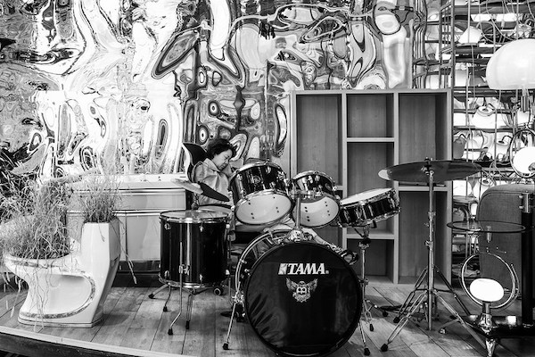 Seated behind a shiny black Tama-brand drum kit on a raised wooden platform, a woman catches a snooze. A psychedelic mural adorns the wall behind her, and there are several pieces of furniture and a white porcelain toilet with plants growing out of it.