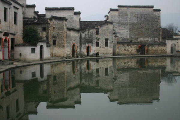 A group of two-story houses with pale gray brick facades, dark roof tiles, and arched doorways—some adorned with bright red lintel scrolls featuring calligraphic couplets—are perfectly reflected in the still water of an adjacent lake.