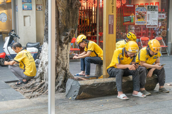 Five food delivery drivers, dressed in bright yellow shirts and bright yellow bicycle and motorcycle helmets, sit or squat near a tree outside a restaurant as they look down at their mobile phones.