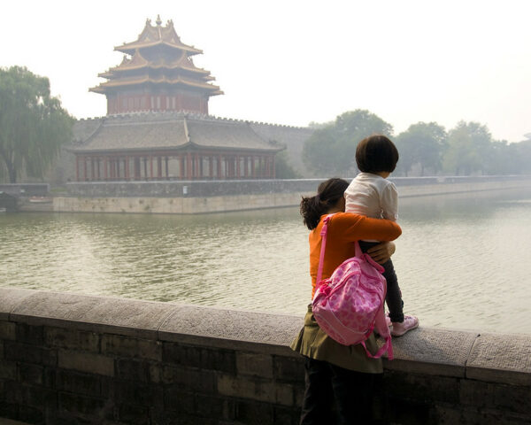 A woman in an orange shirt and pink backpack holds a toddler tightly as the child stands atop a low stone wall looking out at a moat and a corner building of a red-and-gold traditional style Chinese palace.