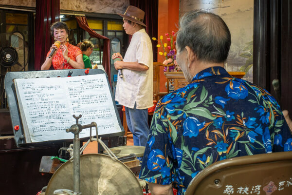 A balding male musician in a Hawaiian shirt looks at his sheet music, while the two performers in front of him (a casually dressed older man in a hat, and an older woman in a red and white qipao) present a Cantonese Opera performance upon a stage decorated with a classical Chinese landscape painting, traditional wooden Chinese furniture, and brightly colored flowers.