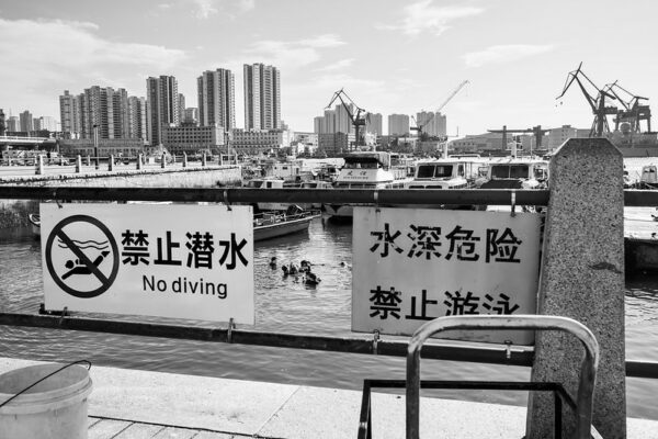 A group of swimmers blithely ignore posted signs warning against swimming or diving in Qingdao’s busy waterfront area. Behind the swimmers are moored fishing boats, and beyond those, towering construction cranes and high-rise apartment buildings.